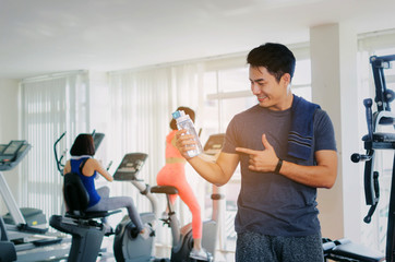 muscular asian young handsome man with towel showing thumbs up and drinking water while resting after workout in fitness gym, bodybuilder, healthy lifestyle, exercise and sport training concept