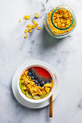 a bowl of smoothies, with cereals for a healthy breakfast and berries, chia seeds and coconut chips freshly sliced fruit, Wheat bran and Cornflake. Served with a wooden spoon on a white marble table