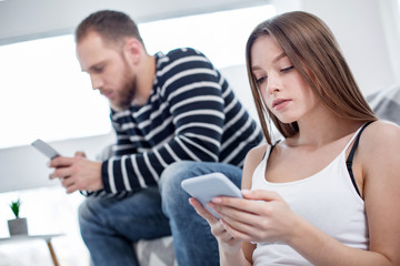 Concentration. Beautiful concentrated long-haired woman holding her phone and her man sitting on the couch and using his phone
