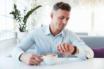 Portrait of handsome man sitting alone in city cafe with cup of cappuccino, looking on wrist watch and waiting for meeting