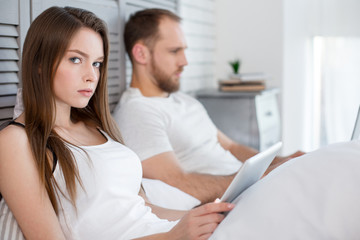 Fototapeta premium Dissatisfaction. Attractive angry long-haired young woman using her tablet and her man lying in bed next to her and paying no attention to her