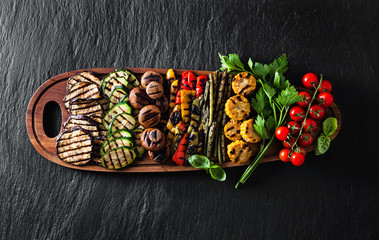 a large portion of colored grilled vegetables and mushrooms on a wooden tray on a dark slate....