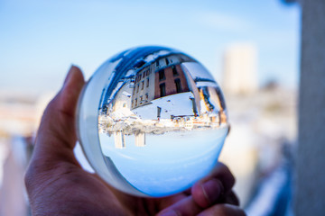 A hand holding a crystal ball for optical illusion. City as the background. Known as an orbuculum, is a crystal or glass ball and common fortune telling object. Performance of clairvoyance and scrying