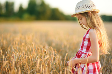 Adorable girl wearing straw hat walking happily in wheat field on warm and sunny summer evening.