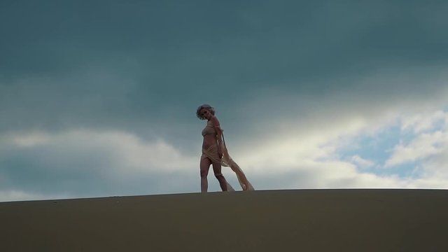 a young woman dressed in urban clothes, a lady posing while shooting fashionable advertising, she walks along the sand in the desert during a ugane or a strong wind