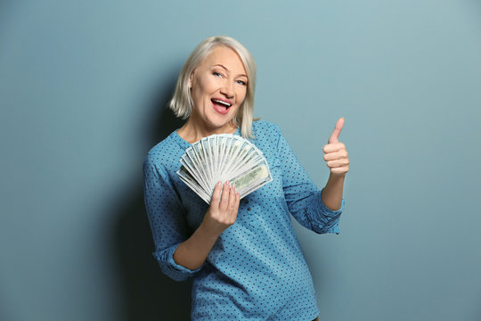 Happy Mature Woman With Money Showing Thumb-up Gesture On Color Background