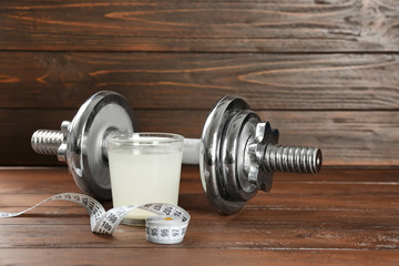 Glass with protein shake, measuring tape and dumbbell on wooden background
