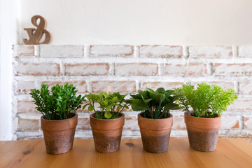 Four artificial plants in clay pot on wooden table with white brick wall textured background in room with copy space.