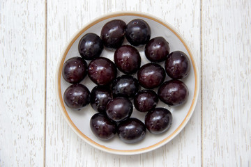 Fresh ripe dark plums on plate, white wooden table, top view