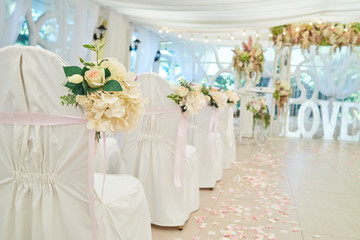Beautiful flower arrangement on white wedding chairs on each side of archway, selective focus. Empty chairs with white cloth for guests on wedding ceremony, free space