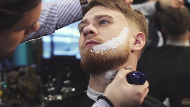 Handsome young man getting his beard shaved at the barbershop professional barber applying shaving cream on his face and neck beardcut barbering service customer fashion professional.