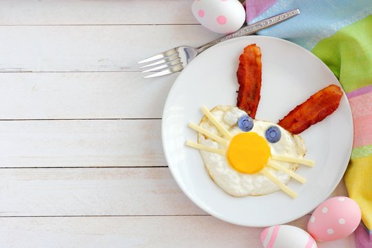 Easter breakfast with cute bunny face made of egg and bacon. Top view scene with decor over a white wood background.