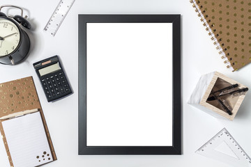 Top view on white desk with black empty mockup photo frame, alarm clock, notebook with golden dots, calculator and marble pail for pens