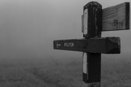 An old sign on the Appalachian Trail points to a shelter written on the wooden board. The background is white with fog and mist. Zero visibility