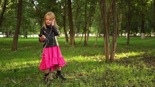 Cute little blonde girl wearing a leather jacket and a pink dress listening to the music standing in a park on a summer day. Locked down real time medium shot