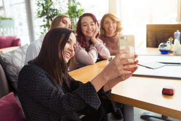 Young beautiful girls taking selfie photo at cafe or coffee shop. Happy women friends having fun, talking together and looking photos at mobile phone. Female friendship, communication concept