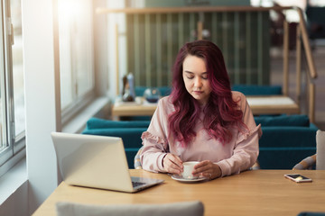 Young hipster girl works at laptop in a cafe, typing on the keyboard. Workplace in a modern sunny cafe, a cup of tea near a laptop