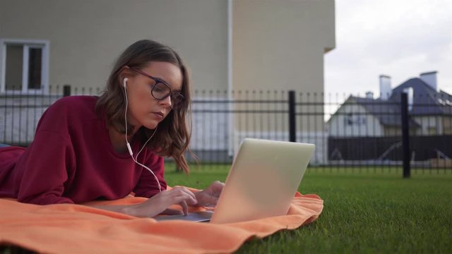 Hipster girl wearing glasses and a red sweater is lying on the grass in her backyard with a laptop, chatting with friends online and listening to the music. Locked down real time medium shot