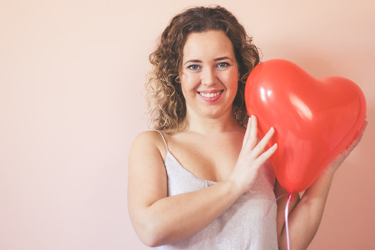 ​Beautiful curly young woman holding a heart shape air balloon on color background. Valentine's Day concept, symbol of love.