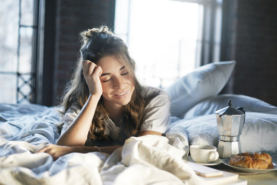 Picture Of Joyful Lazy Young Woman With Messy Hair Lying Awake In Bedroom After Good Sleep, Having Breakfast In Bed Made By Her Husband, Feeling Happy And Relaxed, Going To Drink Coffee With Croissant