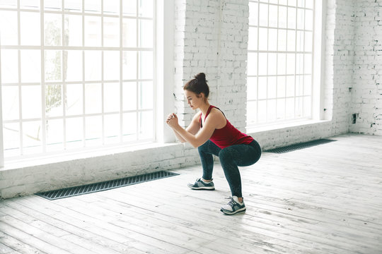 Picture of strong sporty girl wearing stylish tank top, sneakers and leggings doing squats on wooden floor at gym center against large windows background, having determined serious expression