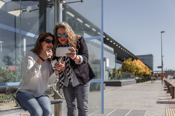 Two businesswomen with casual clothes carefully review a tablet, while they have a coffee and wait at a bus stop