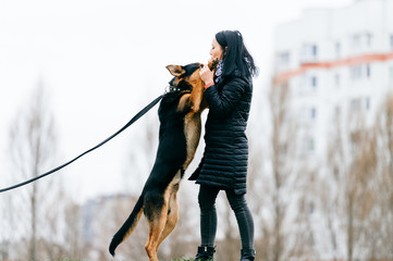 Young active black haired girl in down jacket playing with her lovely dog outdoor. Cheerful happy female have fun and training  puppy with stick. Cute woman dancing with purebred pet. Walk the big dog