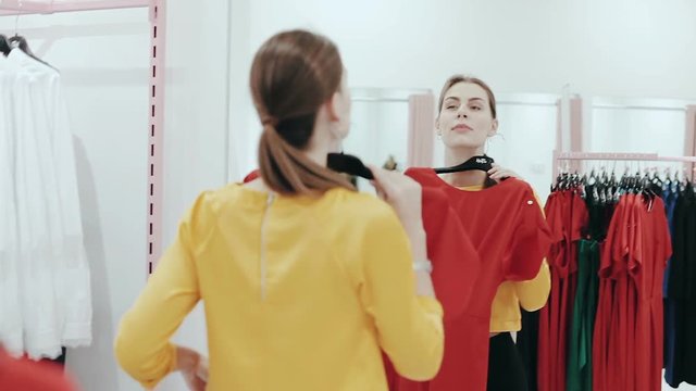 Young slim lady with ponytale choosing a dress in clothes shop. Woman shopping and checking clothes in front of mirror. Style and fashion concept