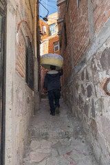 Obraz na płótnie Canvas From the back, a man with a basket of laundry on his head, walking up a narrow stone pathway, between two stone walls, in Guanajuato, Mexico
