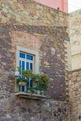 Aged stone wall with a long window, a balcony with some potted plants, and a sliver of pink, in the background, in the old city of Guanajuato, Mexico-a UNESCO Heritage site - 192074424