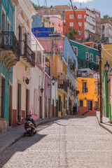 A hilly cobblestone street with many colorful houses and one scooter, parked, in Guanajuato, Mexico