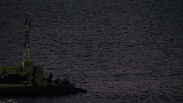 NEA KALLIKRATIA, GREECE - AUGUST 12, 2017: Several men relaxing with fishing from the quay. Evening scene with sea, fishermen and blinking sea-light