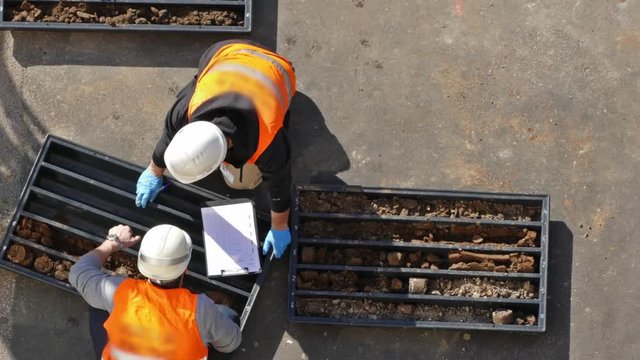 Drilling rig industry. Workers dressed in helmet and working clothes examining soil samples taken from rotary pipe and sorting inside black plastic box for further explorations