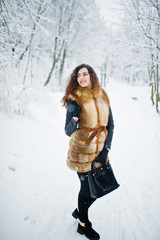 Elegance curly girl in fur coat and handbag at snowy forest park at winter.