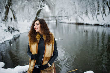 Elegance curly girl in fur coat at snowy forest park agasinst frozen river at winter.