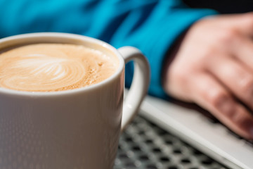 Coffee latte on a table from a cafe, with foam while a person works on a laptop computer