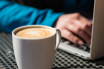 Coffee latte on a table from a cafe, with foam while a person works on a laptop computer