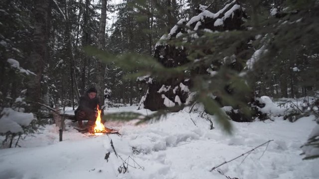Young caucasian guy sitting in forest in front of bonfire tries to warm up on cold winter evening, slow motion