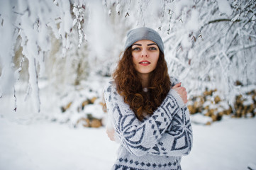 Cute curly girl in sweater and headwear at snowy forest park at winter.
