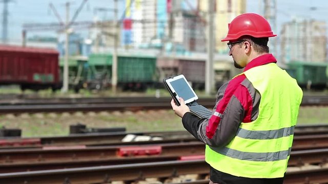 Railwayman with computer at freight train terminal.Railwayman in uniform and red hard hat works with tablet computer. Railway employee makes notes in his tablet pc