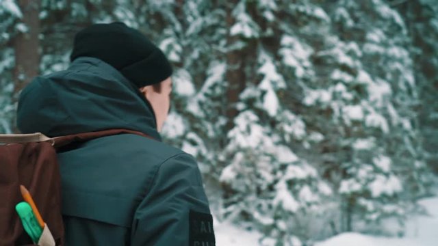 Traveler guy in warm clothes and backpack walking on snow covered trail in pine forest on cold cloudy winter day, slow motion