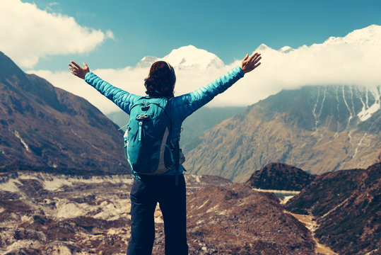 Standing young woman with backpack and raised up arms on the hill and looking on mountains. Landscape with happy girl, mountains, blue sky with clouds in autumn in Nepal. Travel. Trekking. Vintage