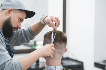 Fototapeta premium Portrait of a hairdresser with a beard who makes a hairdo to a young man. Creation of stylish men's hair scissors in the men's beauty salon. Haircut in barbershop