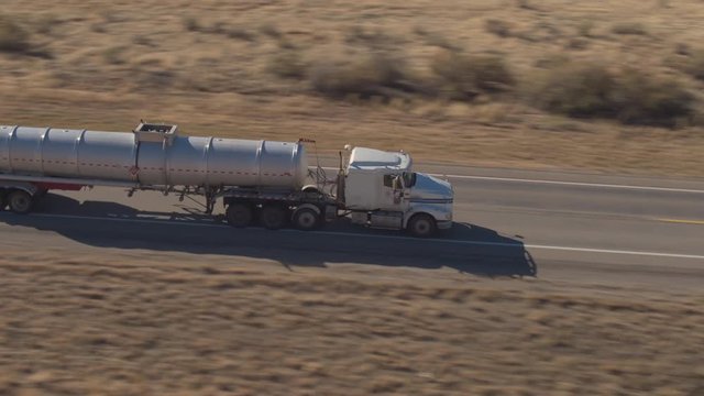 AERIAL CLOSE UP: Unbranded white semi truck driving on empty road, transporting goods on sunny day. Freight container truck hauling and delivering cargo across a country. Trucking logistics shipping