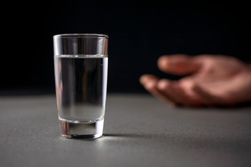 glass of vodka or alcohol drink, hand of a drunk man in the background, alcoholism and alcohol abuse concept, defocused, selective focus, close up, gray table, dark background