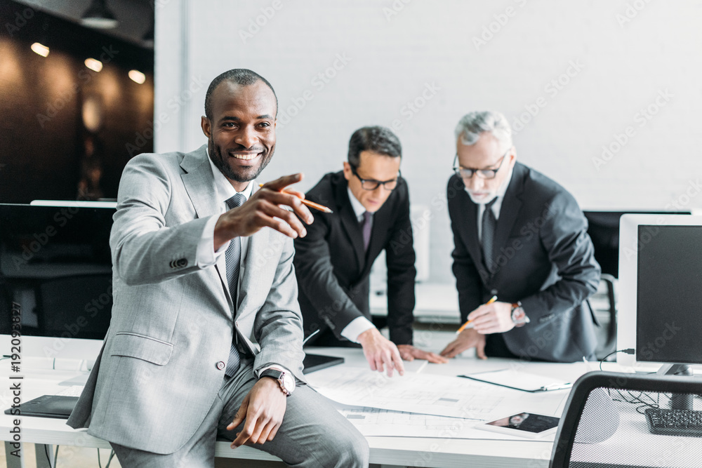 Wall mural smiling african american businessman pointing away while business colleagues discussing work in office