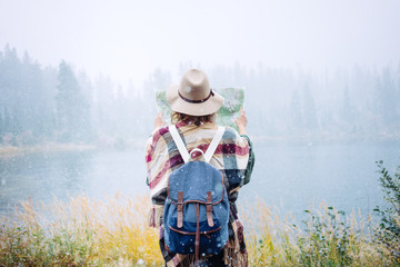 Travel woman reading map among amazing  wilderness, enjoy the nature landsape. Forest and lake, wearing hat and poncho, boho and wanderlust style