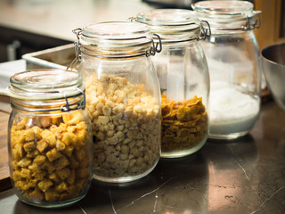 Glass bottles with cereals, crispy dice breads, chocolate chips, flour and icing sugar in a row on wooden table in kitchen or pantry for topping or ingredients of dessert or breakfast