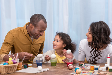 african american parents and daughter painting easter eggs and holding them above glasses with paints