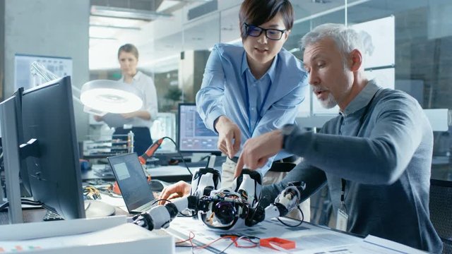 Two Engineers Talking while Working with Robot, Manipulating its Responses and Checking Data with Laptop Computer. In the Background Robotics Research Center Laboratory with Specialists Working.